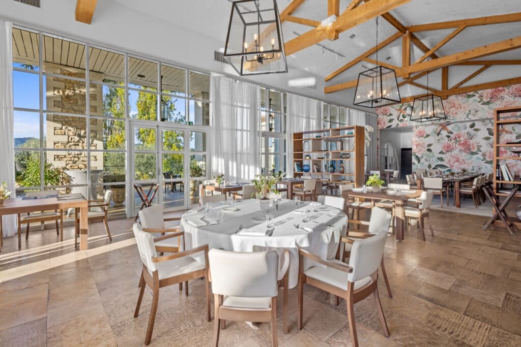 Restaurant lumineux : poutres en bois, papier peint fleuri, grandes fenêtres, tables rondes aux nappes blanches, chaises.