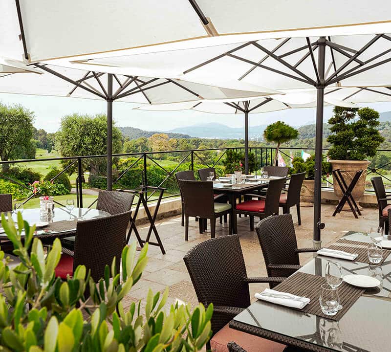Terrasse extérieure avec chaises en osier et tables dressées sous grands parasols blancs. Paysage pittoresque.
