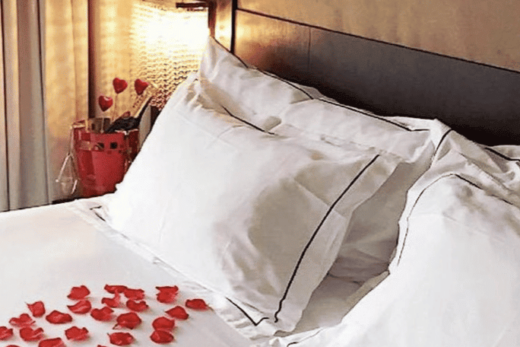 Lit avec draps blancs, pétales de roses. Lampe de chevet et rideau en arrière-plan.