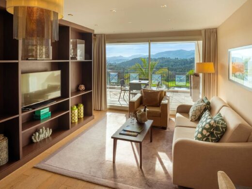 Salon moderne avec canapé beige, table basse en bois ; balcon avec vue montagnes et verdure.