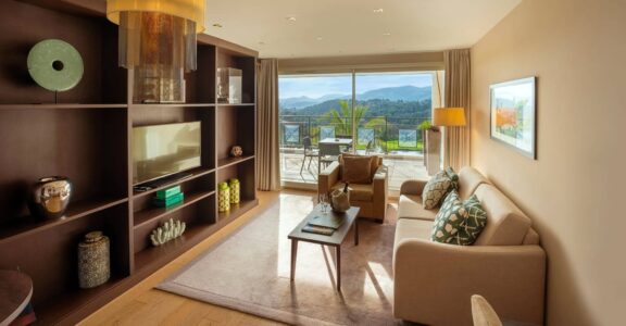 Salon moderne avec canapé beige, table basse en bois ; balcon avec vue montagnes et verdure.
