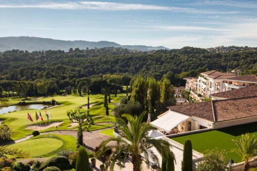 Vue aérienne d'un parcours de golf avec club-house, collines et maisons sous un ciel dégagé.