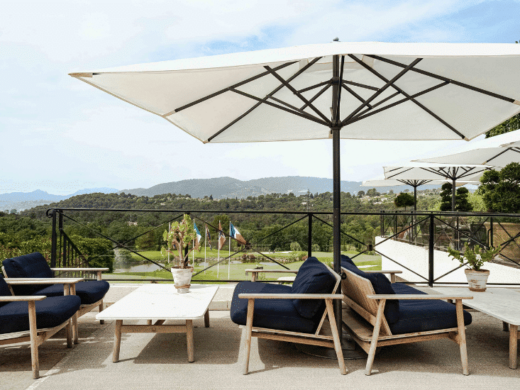 Chaises en bois bleues, tables, parasols blancs, vue sur paysage pittoresque avec collines verdoyantes.
