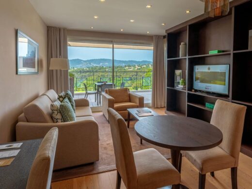 Salon moderne : table ronde, canapé beige, TV, étagères. Porte-fenêtre menant à balcon avec vue sur collines verdoyantes.