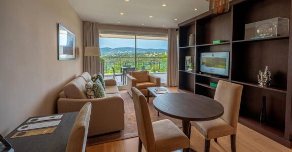 Salon moderne : table ronde, canapé beige, TV, étagères. Porte-fenêtre menant à balcon avec vue sur collines verdoyantes.
