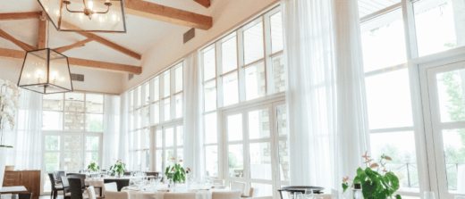 Salle à manger lumineuse avec tables dressées, grandes fenêtres et plantes vertes.