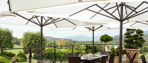 Terrasse extérieure avec chaises en osier et tables dressées sous grands parasols blancs. Paysage pittoresque.