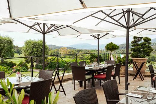 Terrasse extérieure avec chaises en osier et tables dressées sous grands parasols blancs. Paysage pittoresque.