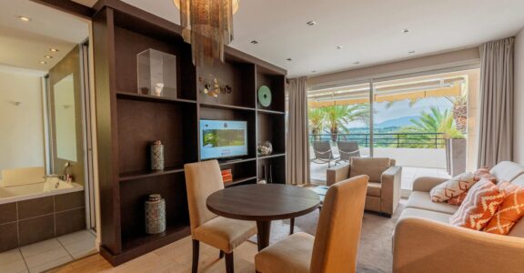 Salon moderne avec table ronde, accès balcon, vue sur mer et palmiers.