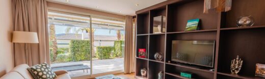 Salon moderne : canapé beige, table basse, grande étagère avec TV, vue sur patio et verdure.