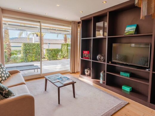 Salon moderne : canapé beige, table basse, grande étagère avec TV, vue sur patio et verdure.