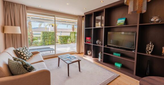 Salon moderne : canapé beige, table basse, grande étagère avec TV, vue sur patio et verdure.