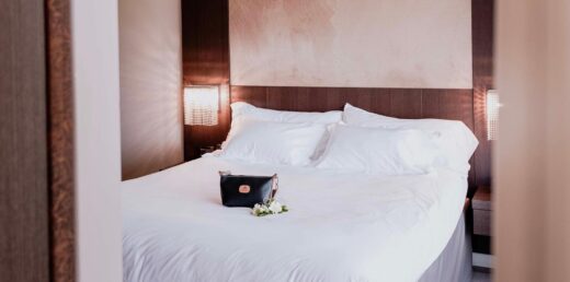 Lit avec draps blancs, lampes allumées à côté, sac à main et fleurs sur le dessus.