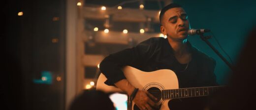 Homme chantant sur scène faiblement éclairée, guitares et lumières en arrière-plan.