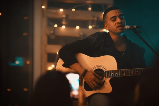 Homme joue guitare acoustique et chante sur scène faiblement éclairée avec guirlandes lumineuses en arrière-plan.