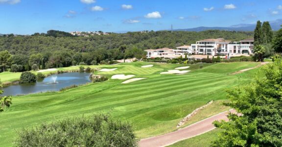 Golf verdoyant, bunkers de sable, eau et bâtiments modernes sous un ciel partiellement nuageux.